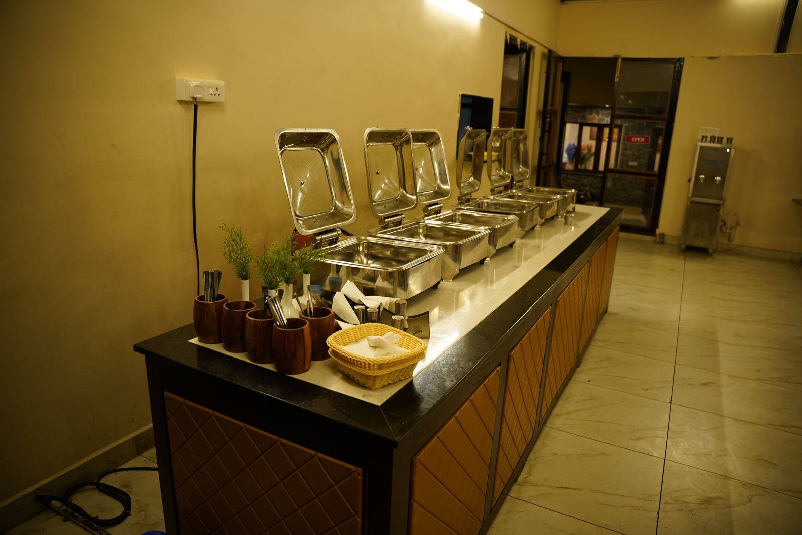 A row of chafing dishes laid out for breakfast service, with cutlery in wooden holders and a bread basket on the counter.