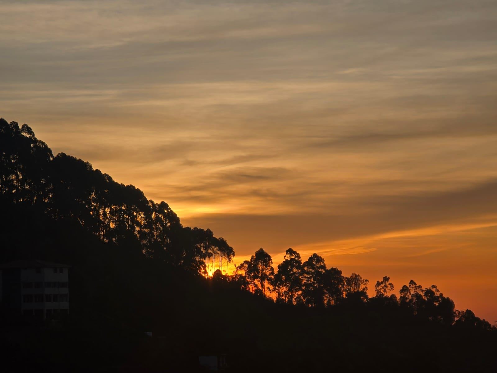 The sun dipping behind a ridge of tall silhouetted trees, its last light spreading orange across a bank of clouds above a hillside building.