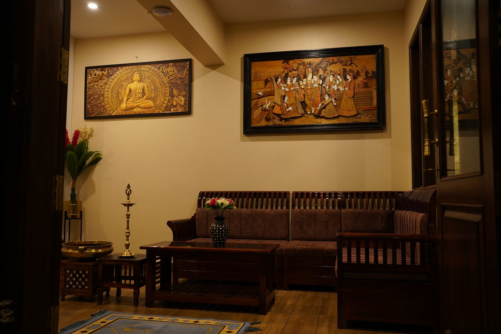 A quiet lounge corner with a dark wood sofa, patterned artwork on the walls, and a brass oil lamp beside fresh flowers.