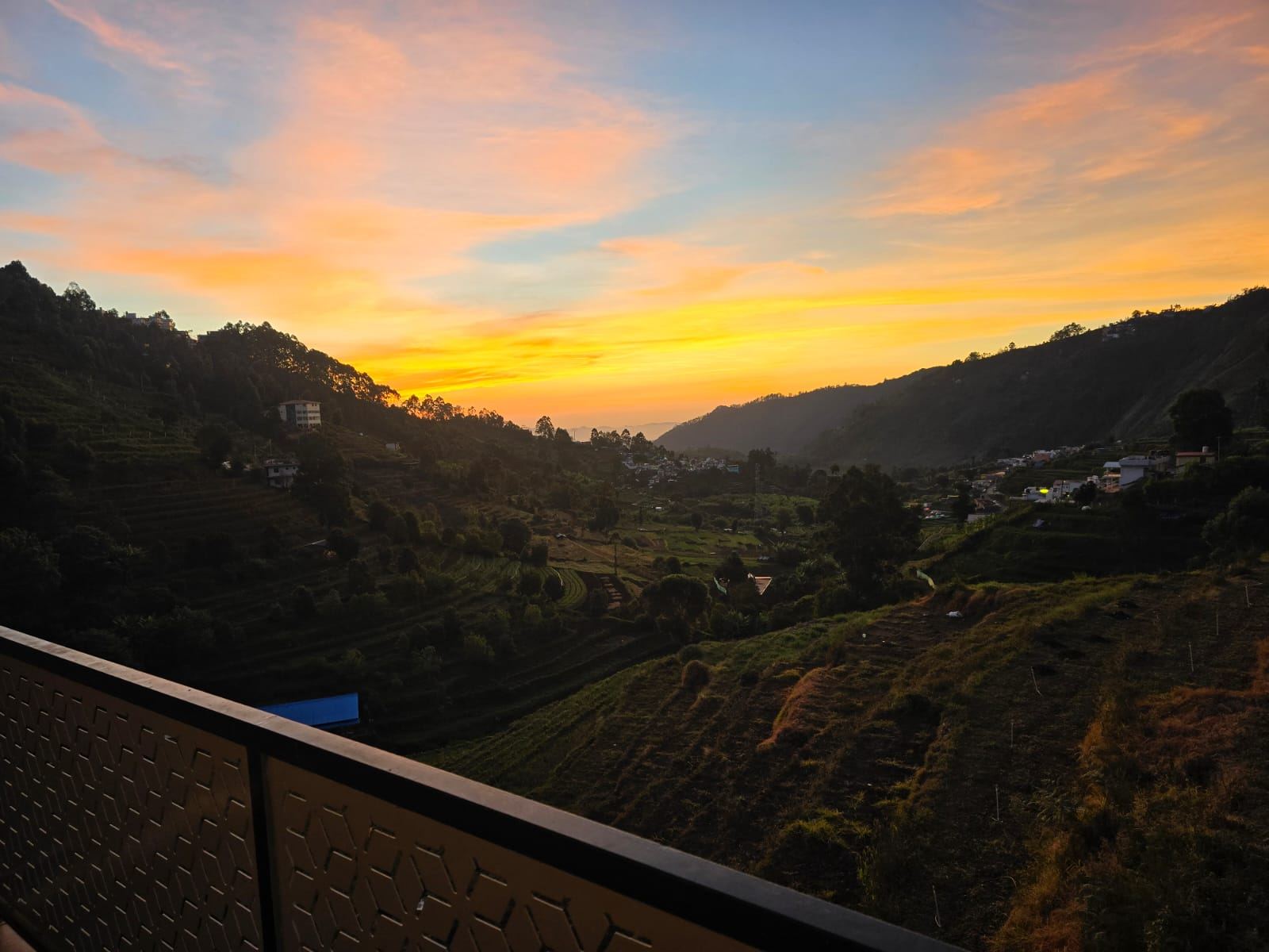A balcony railing in the foreground looking out over terraced hillside fields as the sky turns gold and pink at sunset, village rooftops scattered across the slopes.