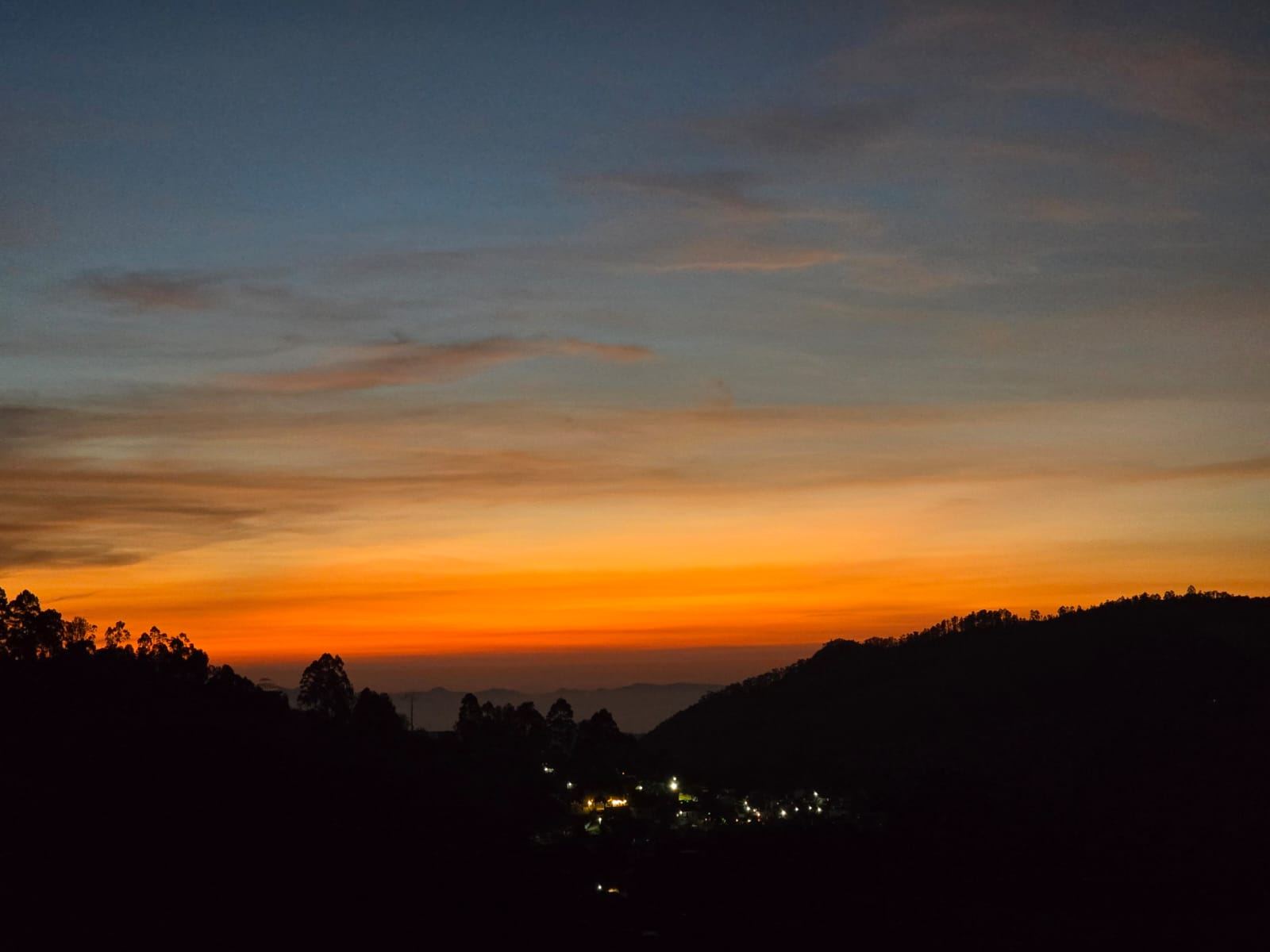 A deep orange band of afterglow along the horizon at dusk, hillside villages lighting up in the valley below dark silhouetted ridgelines.