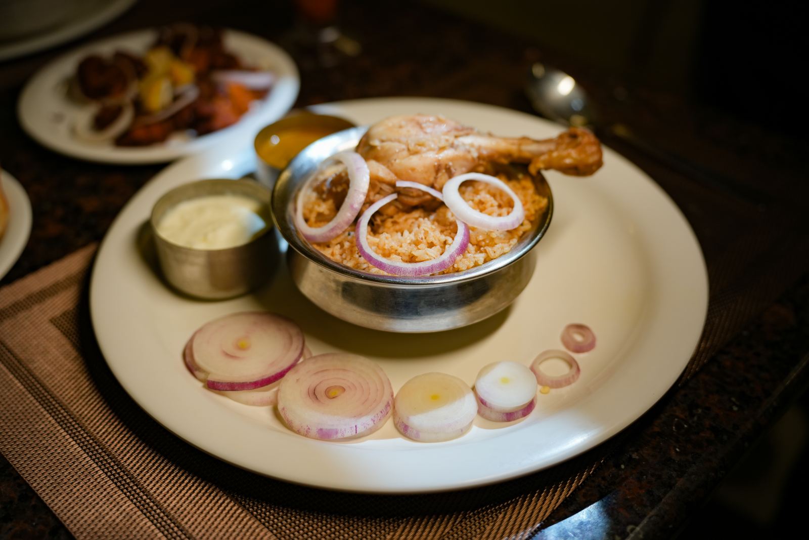 A roasted chicken leg over spiced rice, garnished with onion rings, served with a side of raita