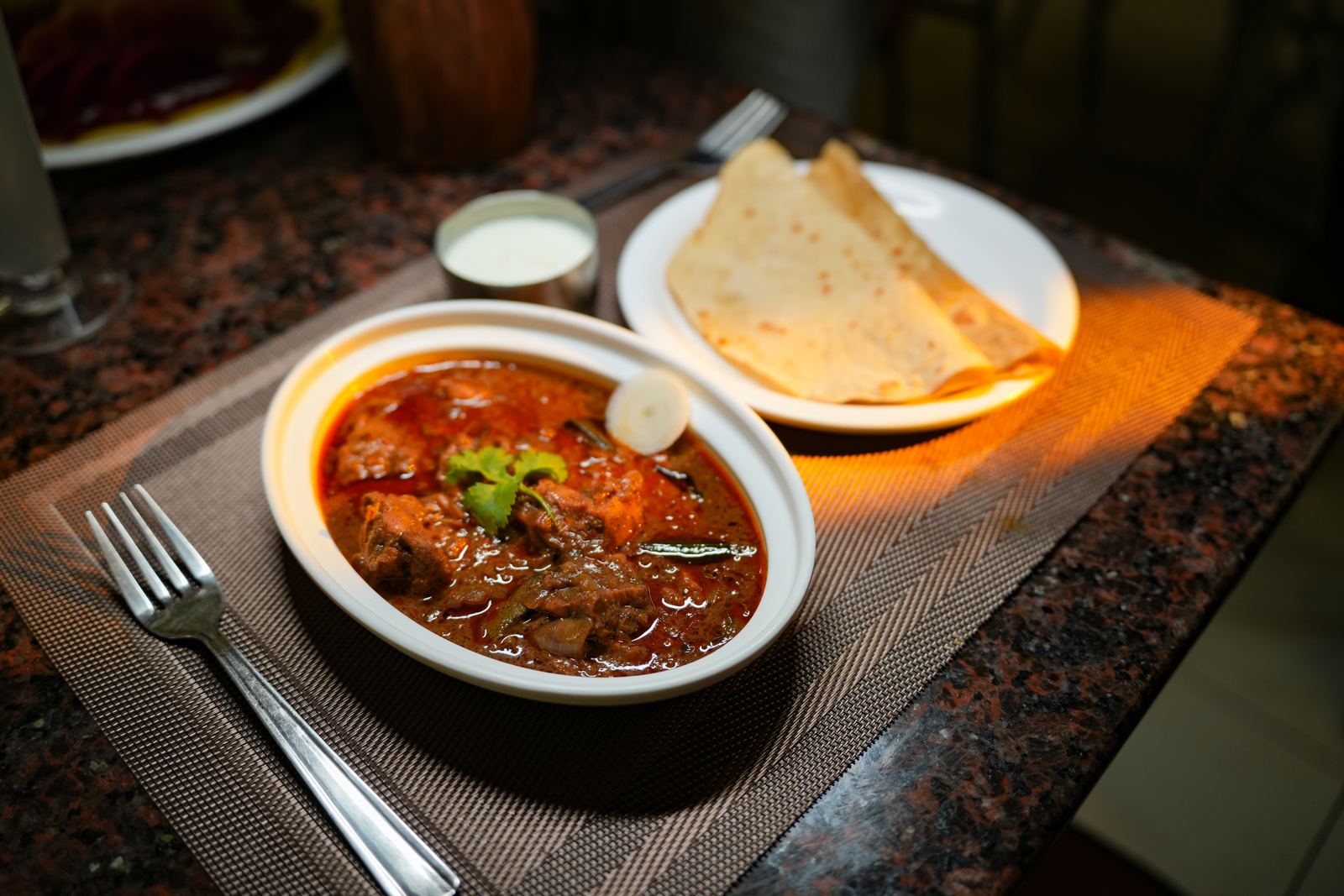 A bowl of spiced meat curry garnished with coriander, served with parathas and a side of raita